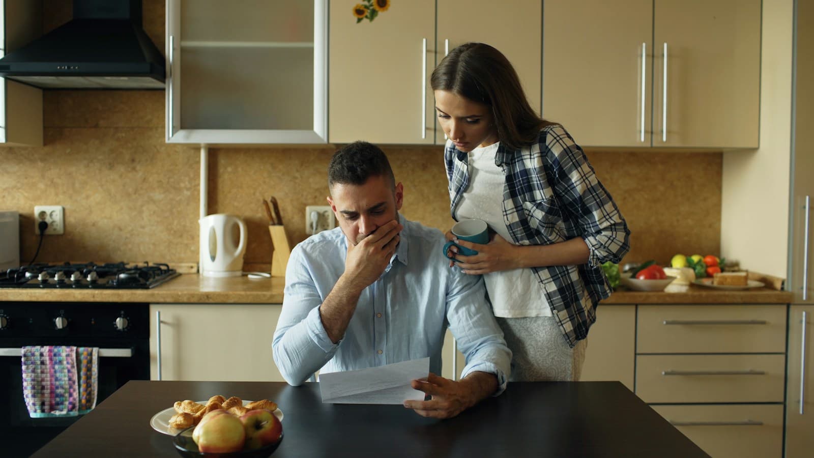 A man reads a bill at a kitchen table while a woman stands beside him holding a mug, both looking at the page.