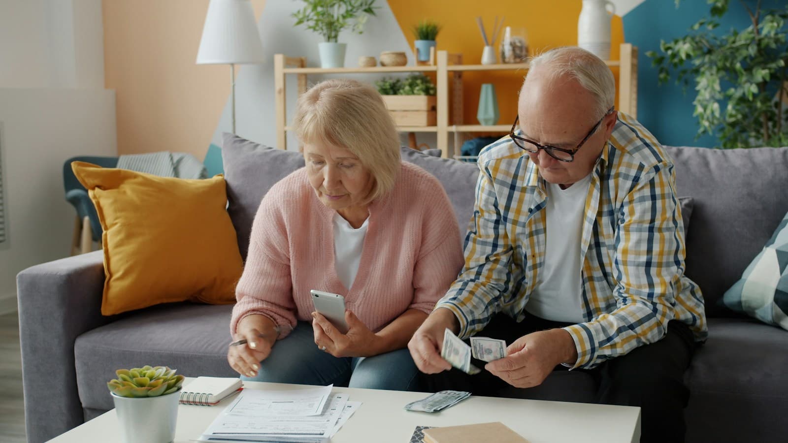 An older couple sit on a sofa with paperwork on the table in front of them, one counting banknotes and the other checking a phone.