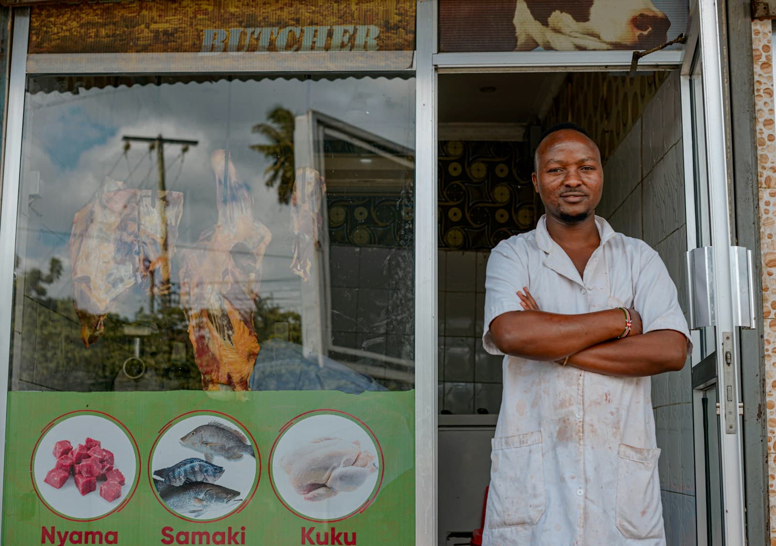 A shopkeeper in a white coat stands with folded arms in the doorway of his butcher shop.